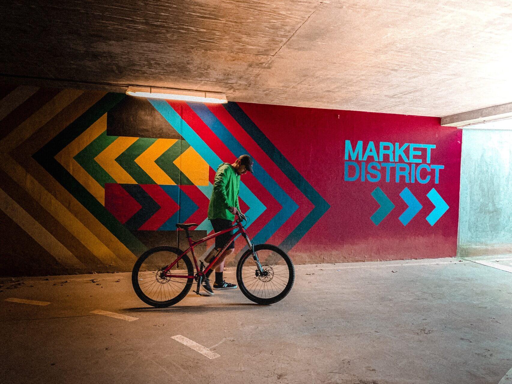 Man standing next to his bike in front of a wall painted red with the text "MARKET DISTRICT" painted in light blue