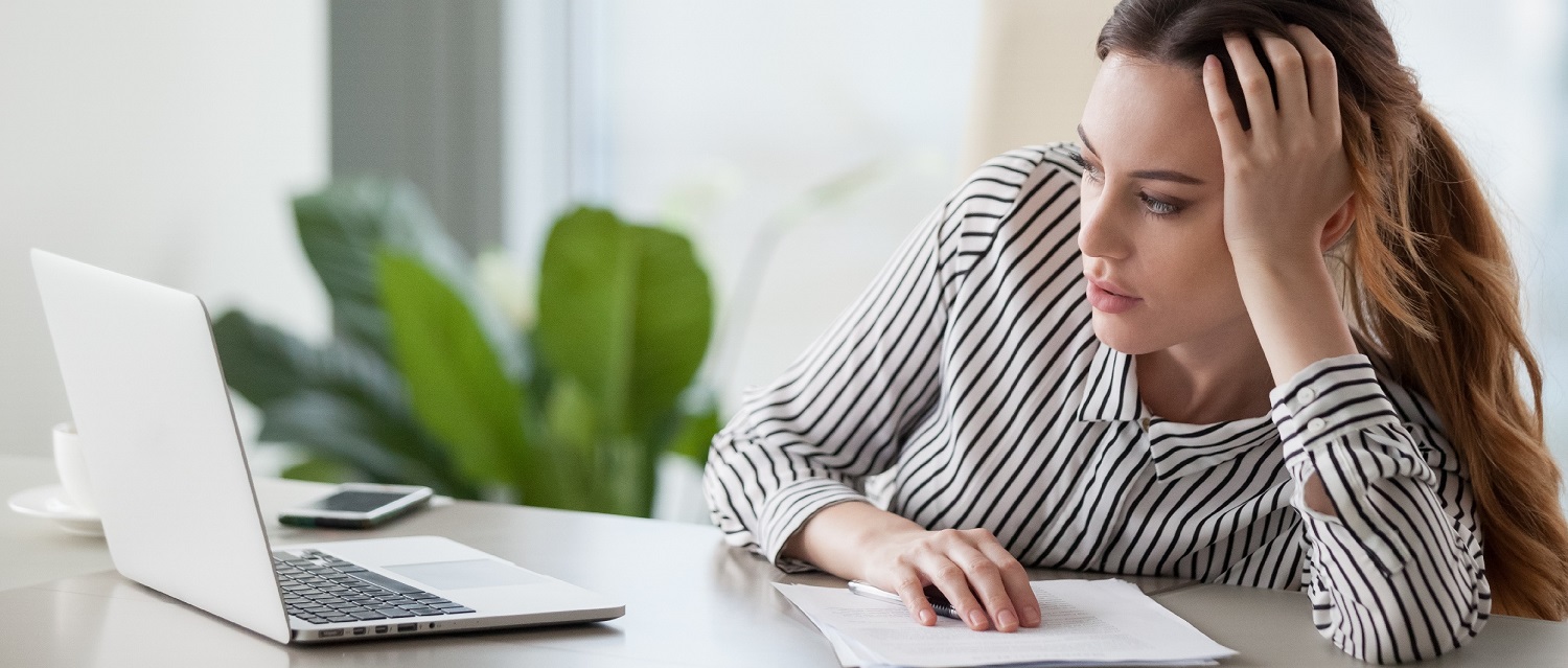 Red haired woman leaning on her left arm looking exacerbated at a laptop on a white table